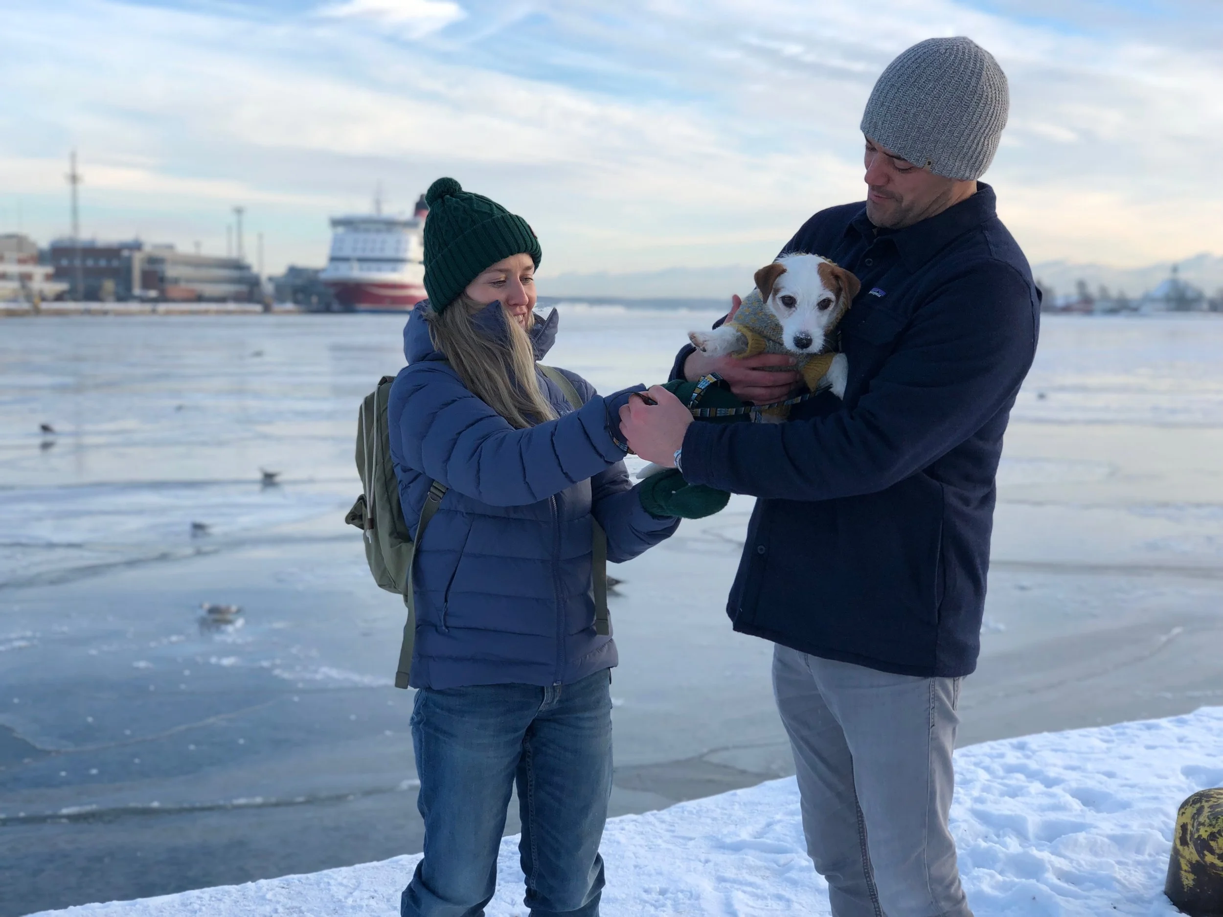A man and a woman with a puppy on a snowy waterfront with boats and buildings in the background.