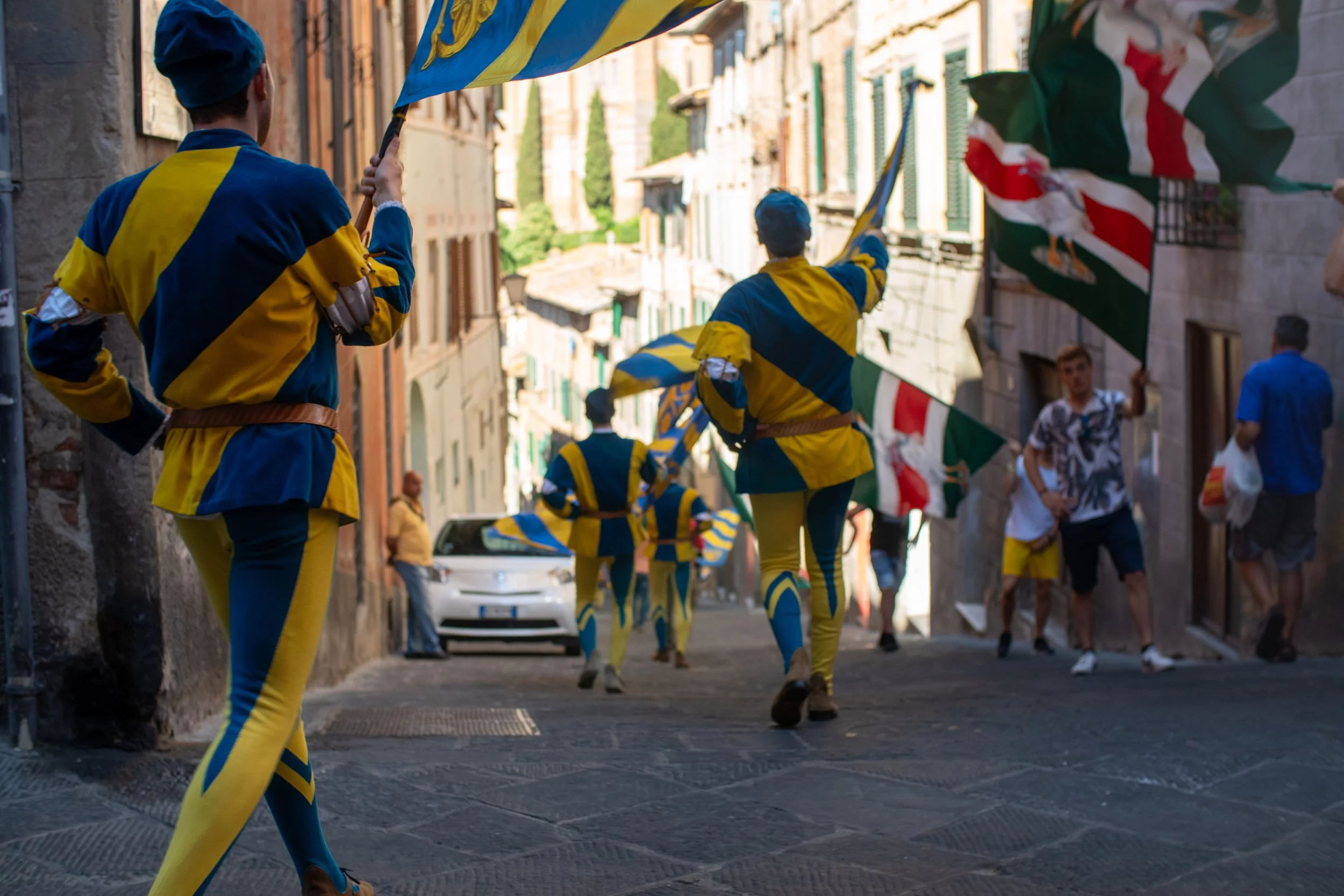 People dressed in yellow and blue costumes performing a traditional parade or dance on a narrow city street, with some holding flags.