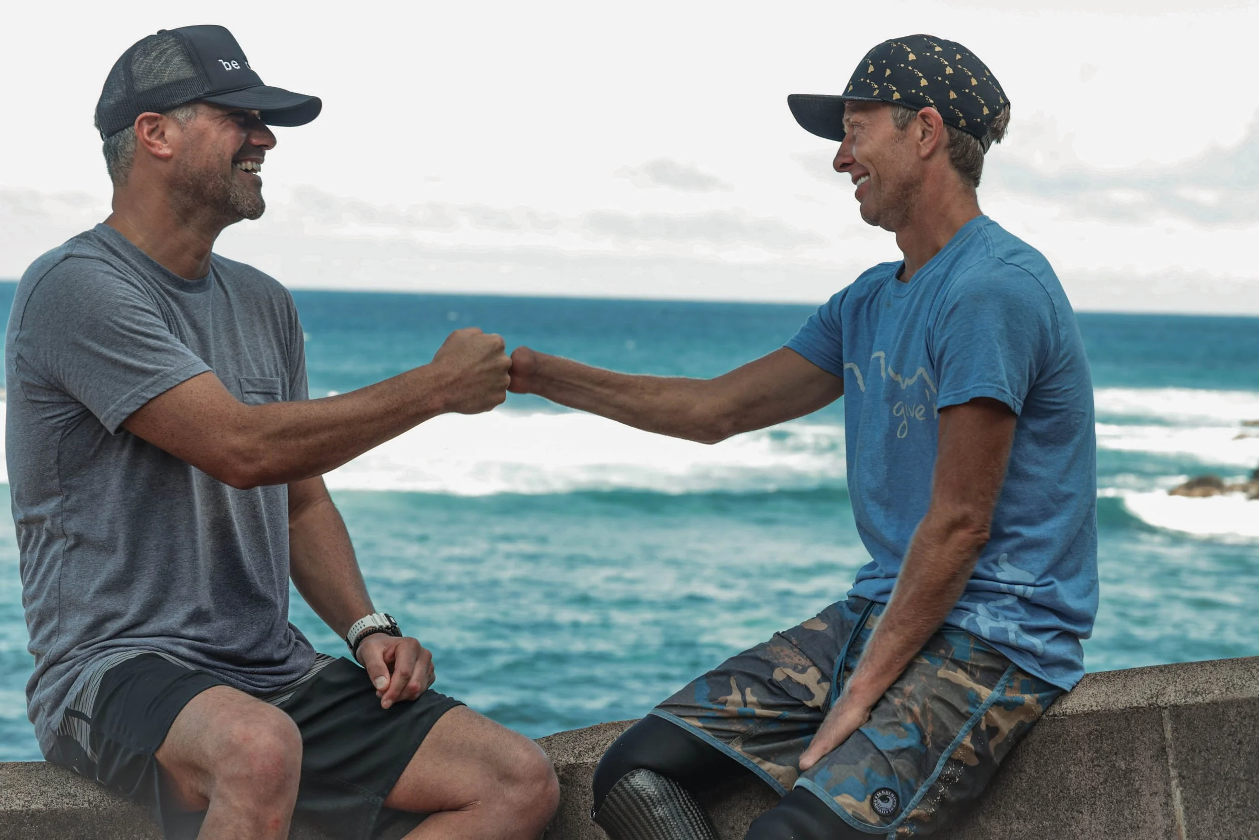 Two men sitting on a concrete ledge by the ocean, smiling and exchanging a fist bump. One man has gray hair, a beard, wearing a gray T-shirt and a baseball cap. The other man has light brown hair, wearing a blue T-shirt, camouflage shorts, and a baseball cap, facing each other with a background of water and cloudy sky.