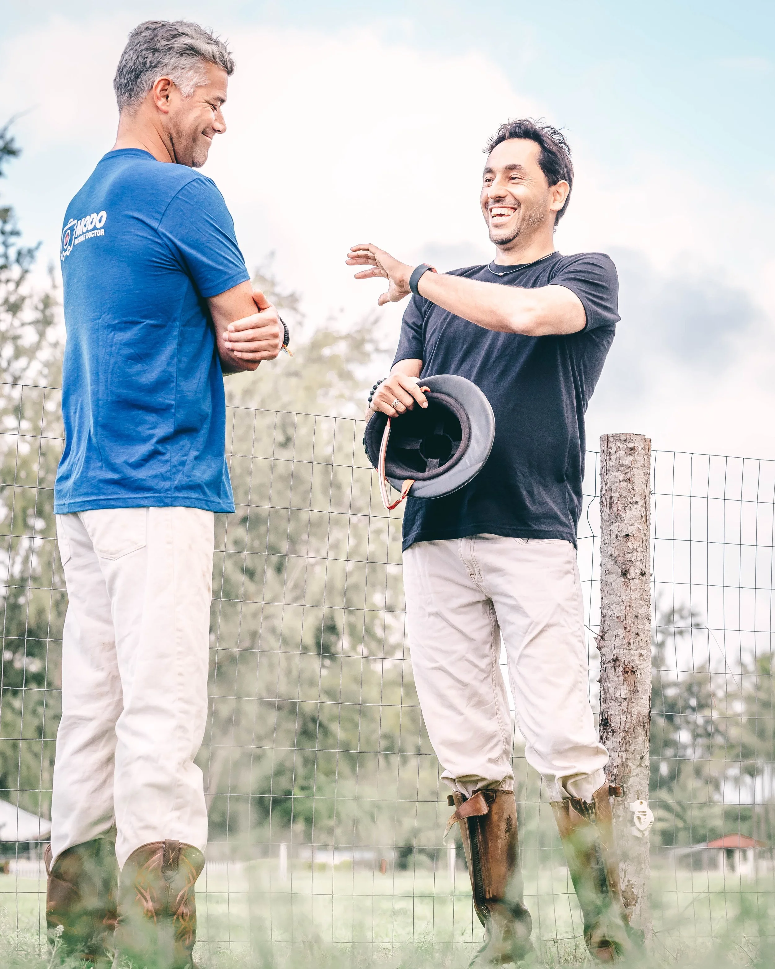 Two men are standing outdoors, smiling and laughing, engaged in conversation. One man is wearing a blue shirt with some writing on the back, beige pants, and brown boots. The other man is dressed in black, with tan pants, holding a black helmet in one hand and gesturing with the other. They are standing by a wire fence with a tree and a cloudy sky in the background.