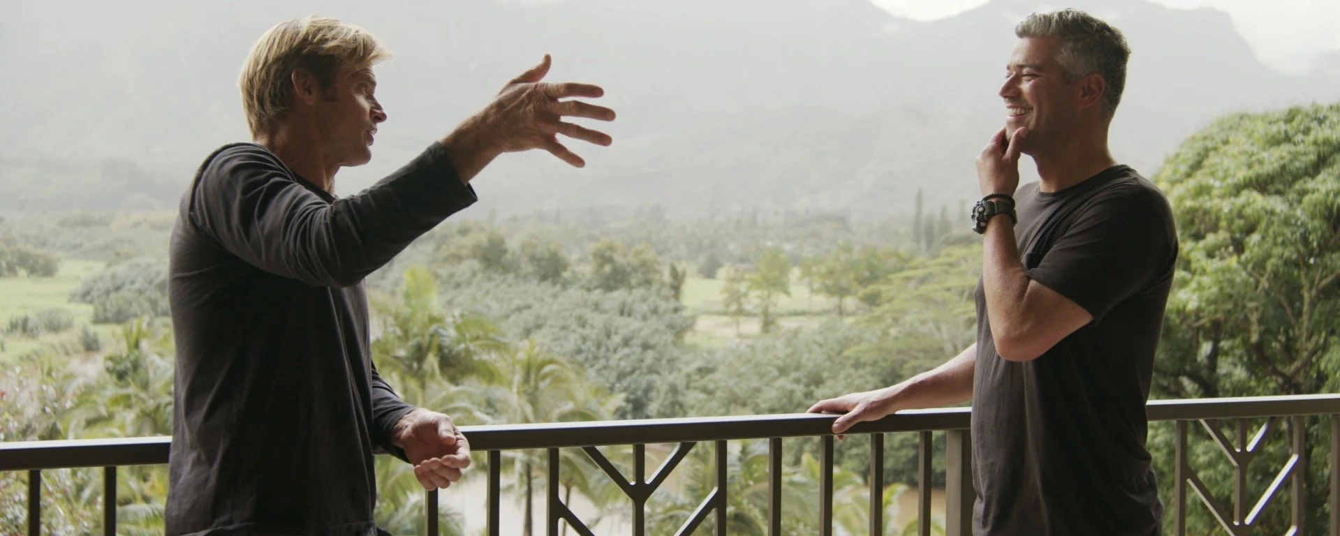 Two men talking on a balcony overlooking a lush, green landscape with trees and rolling hills, engaged in a lively conversation, both smiling.