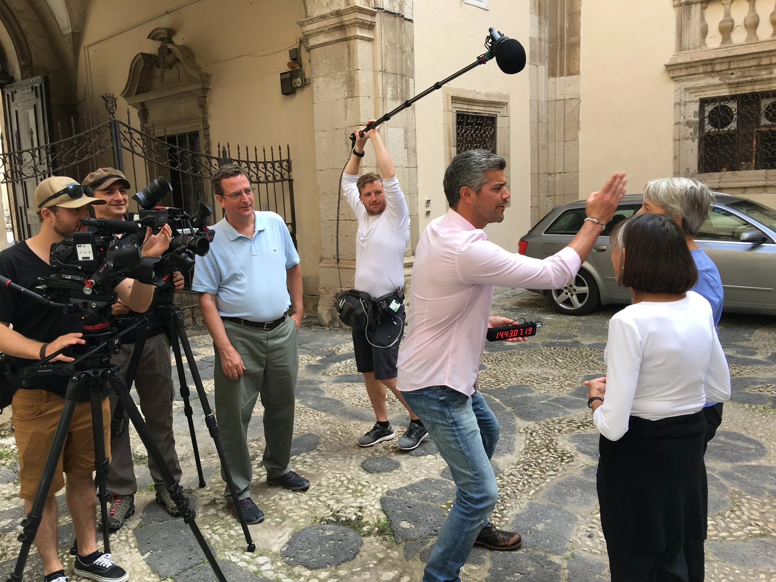 A film crew filming a man in a light pink shirt conducting an interview in an outdoor courtyard with old stone buildings. The crew includes a camera operator, a boom microphone operator, and other crew members, some observing and one preparing to record.