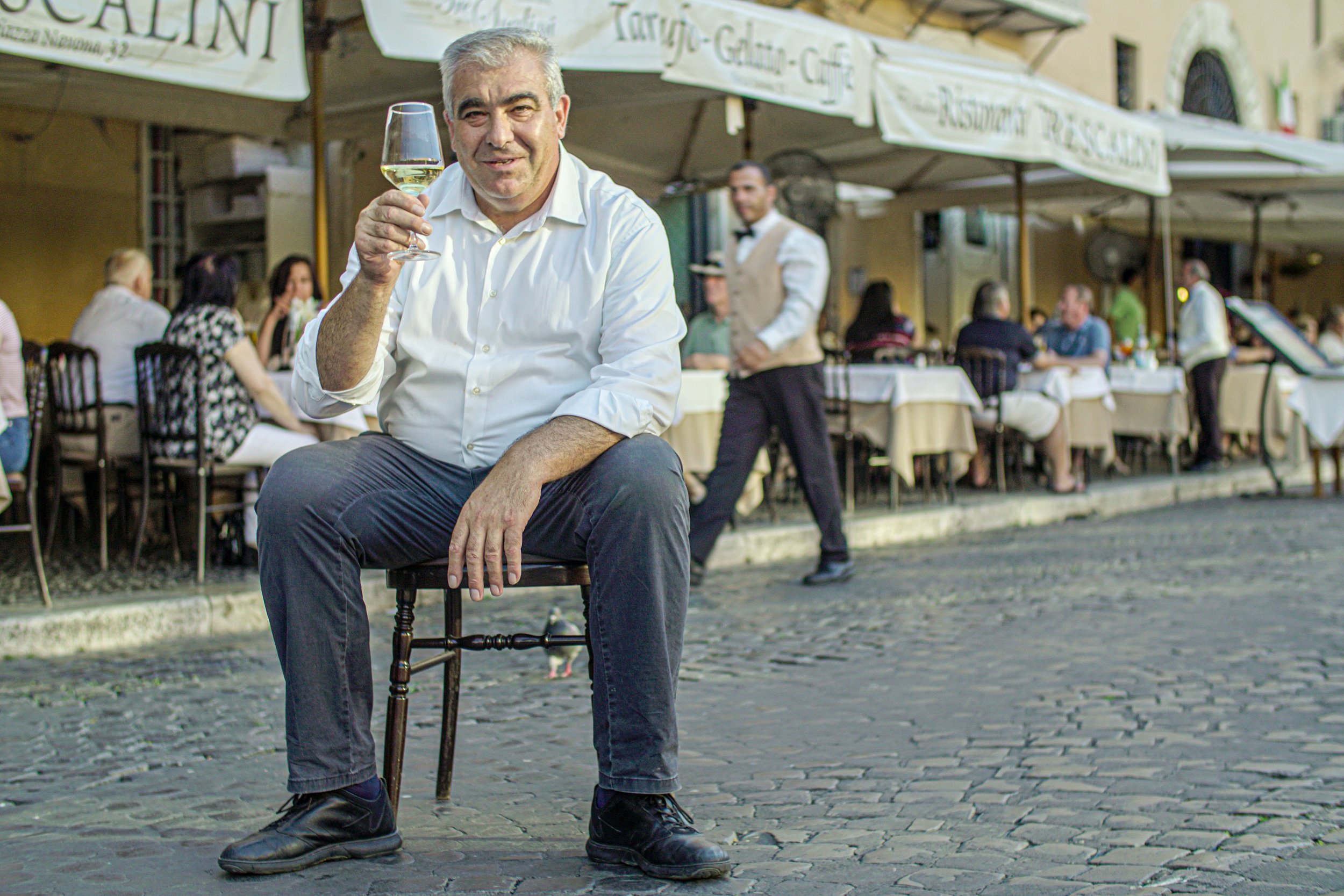 A man sitting on a stool outdoors at a restaurant or cafe, holding a glass of white wine and smiling at the camera. There are tables with people dining in the background under large umbrellas.