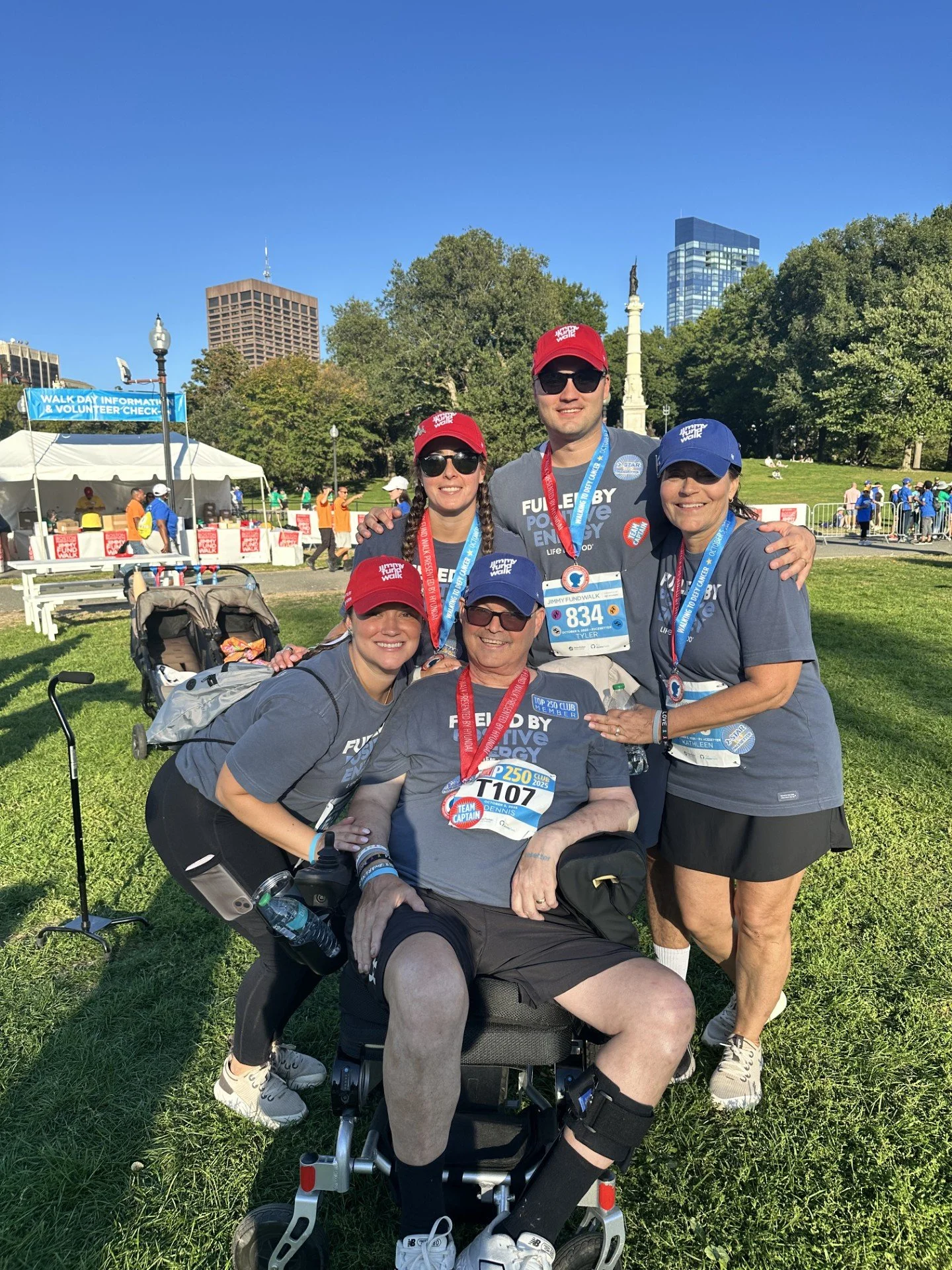 Group of five people at a charity walk event, with some wearing medals, posing outdoors on a sunny day with tall buildings and trees in the background.
