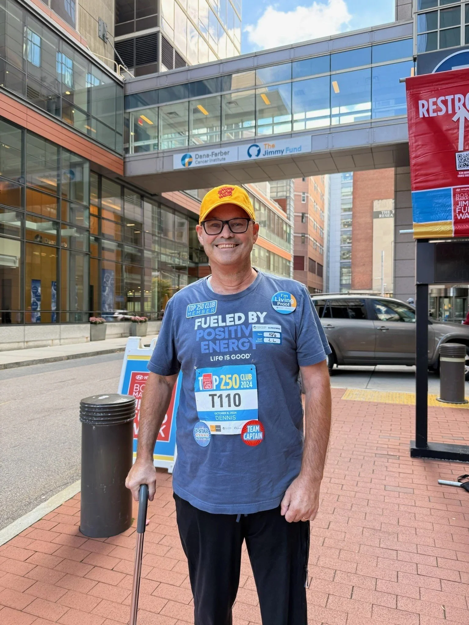 A man wearing glasses and a yellow cap smiling at the camera, standing outside in an urban area with modern glass buildings behind him. He is dressed in a blue shirt with various race and energy-themed buttons and a bib with his name "Dennis" and the number "T110." He holds a cane in his right hand.