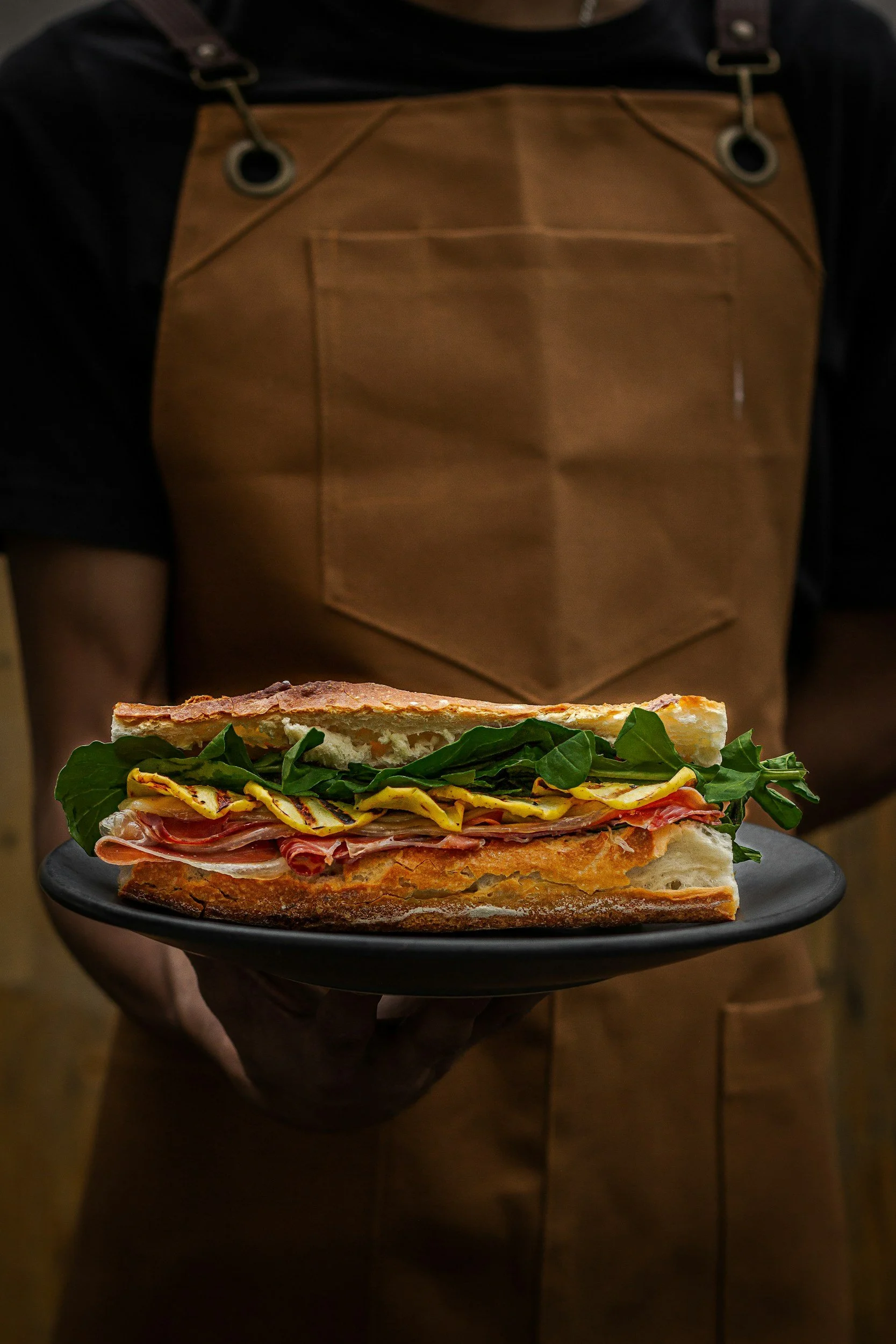 Person wearing a brown apron holding a black plate with a large submarine sandwich filled with lettuce, tomato, meat, and cheese.