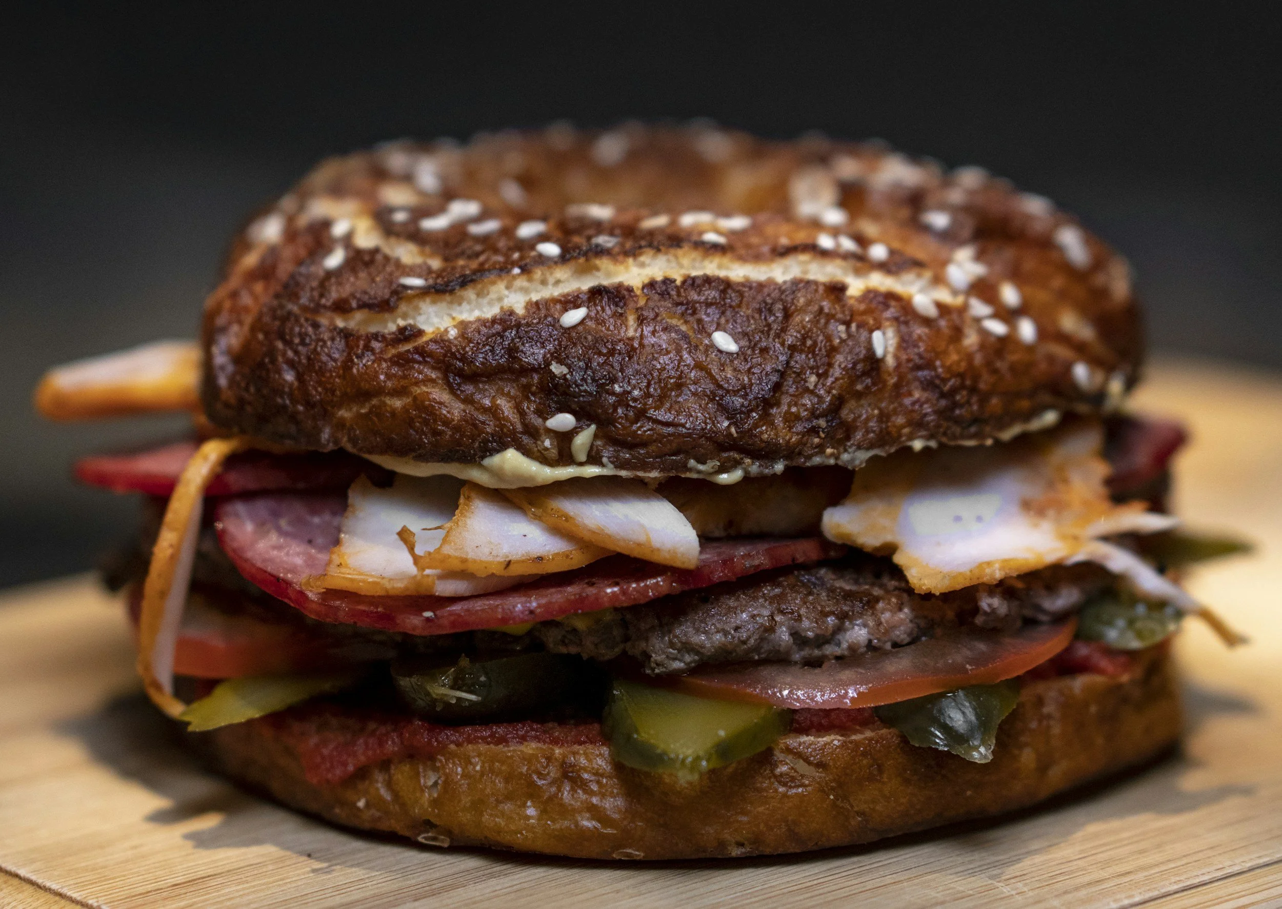 A close-up of a layered burger with a toasted bun, sesame seeds, beef patty, cheese, lettuce, tomato, pickles, and bacon.
