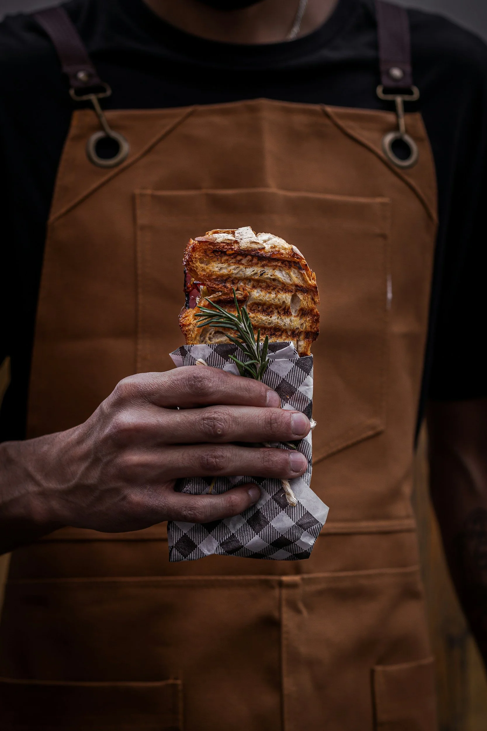 A person wearing a brown apron holding a toasted sandwich wrapped in checkered paper with a sprig of rosemary.