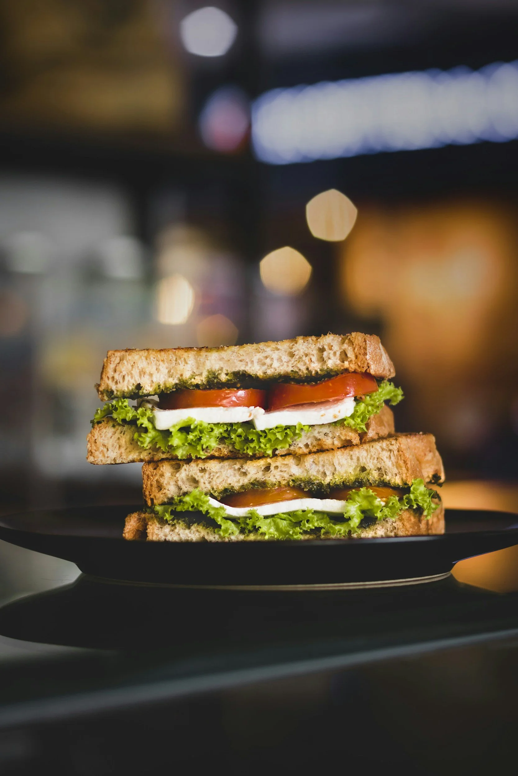 A close-up of a stacked sandwich with lettuce, tomato, and cheese on toasted bread, on a black plate in a softly lit environment with bokeh lights in the background.