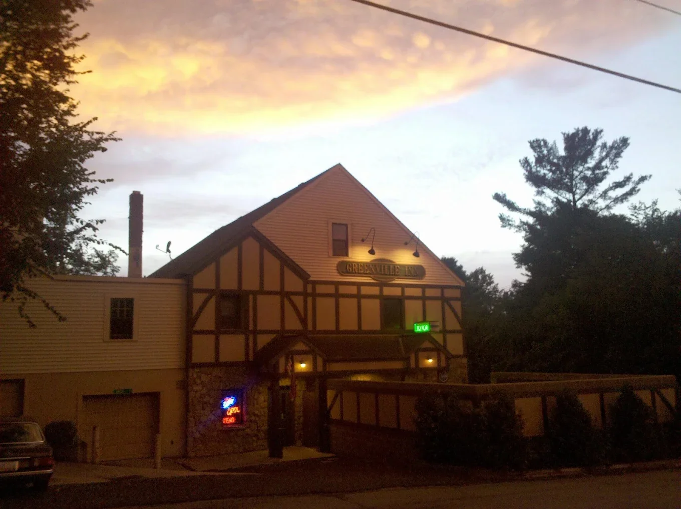 Exterior view of a building with a sign saying 'GREAT VALLEY INN', illuminated signs, trees on either side, a sunset sky with clouds, and a car parked in front.