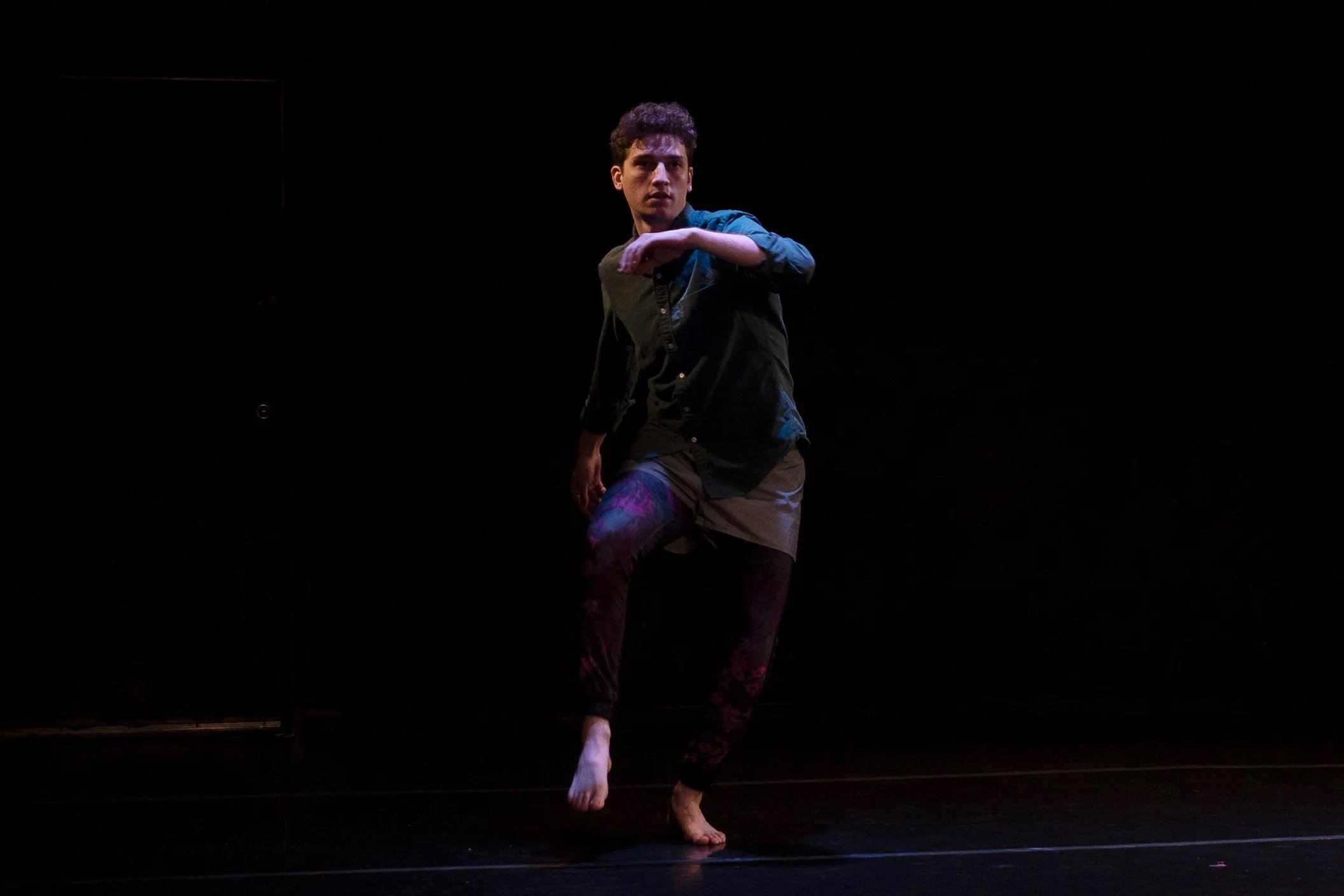 A young male dancer performing on a dark stage, wearing a dark shirt and pants with colorful patterns, in a pose with one arm bent at the elbow and the other arm resting on his knee.
