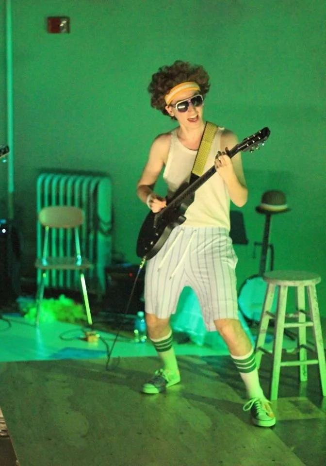 Person with curly hair, sunglasses, and headband playing electric guitar on stage with green lighting, wearing a tank top, striped shorts, and colorful sneakers.