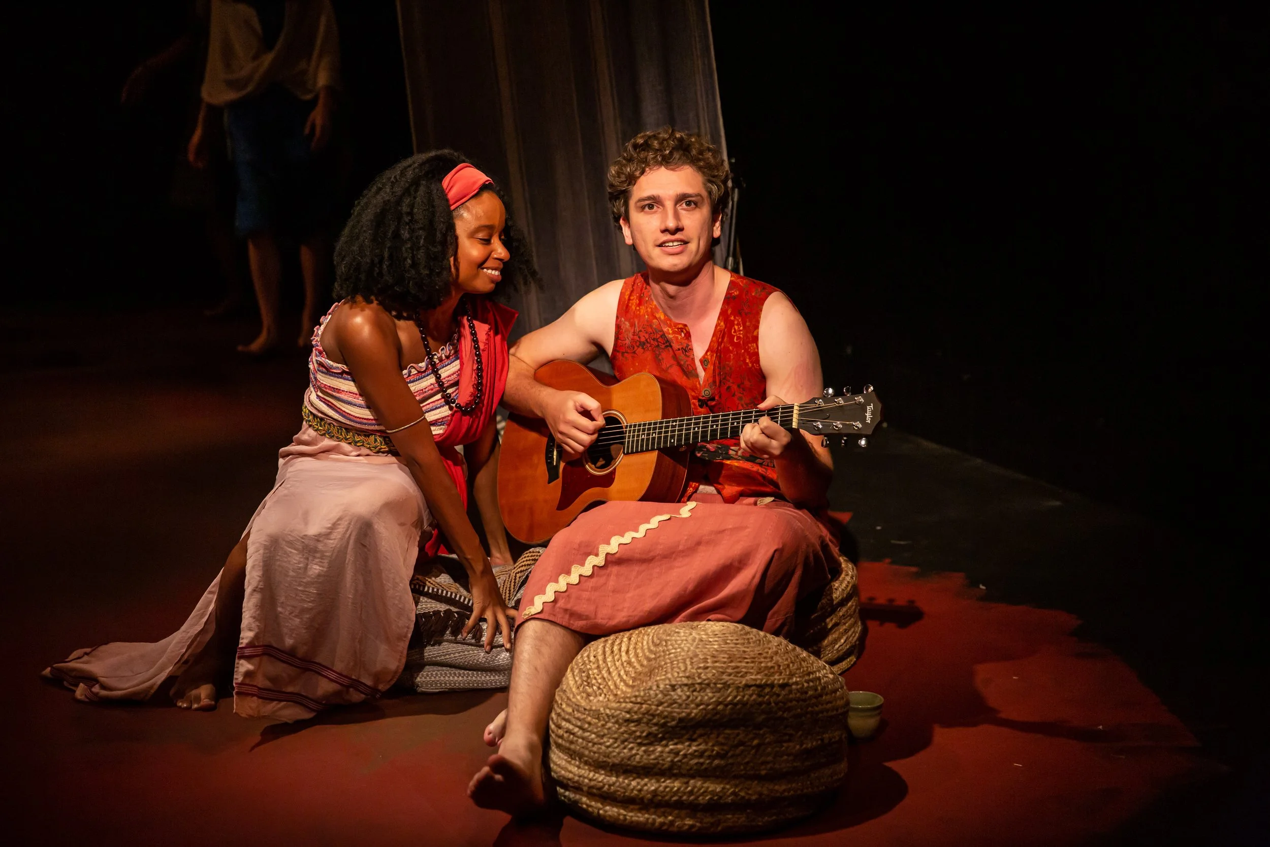 Two performers on stage. The woman is smiling with her eyes closed, wearing a colorful strapless dress and a headband, while the man is singing with an acoustic guitar, wearing a sleeveless orange dress with a floral pattern. They are sitting on wove