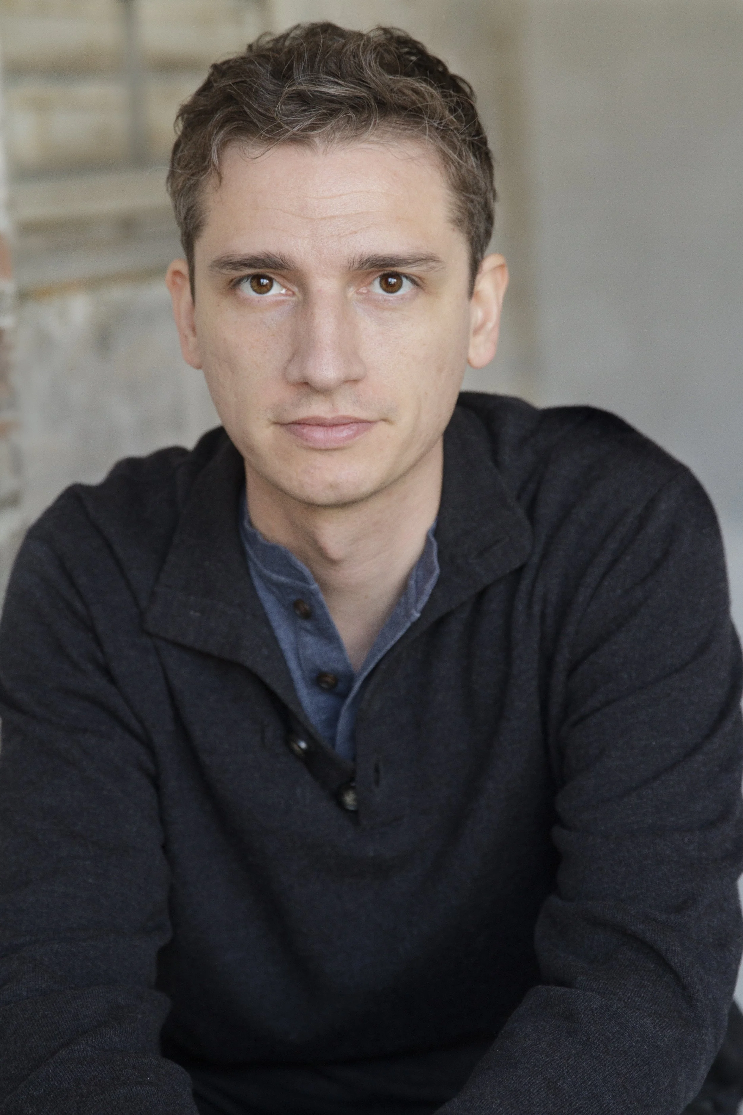 Close-up portrait of a young man with short, curly brown hair, brown eyes, and light skin, wearing a dark sweater over a collared shirt, sitting indoors.