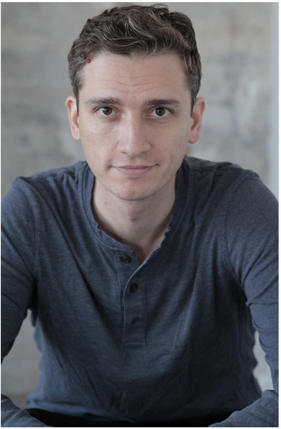 A young man with short brown hair and fair skin, wearing a dark blue henley shirt, looking directly at the camera with a neutral expression, in a room with a blurred concrete wall background.