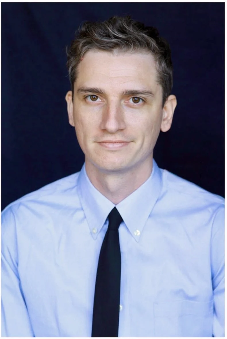 A young man with short, dark, wavy hair wearing a light blue dress shirt and a black tie, standing against a dark background.