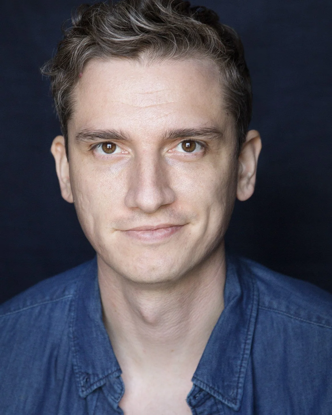Close-up portrait of a man with short brown hair, brown eyes, light skin, wearing a dark blue denim shirt, smiling slightly, against a dark background.