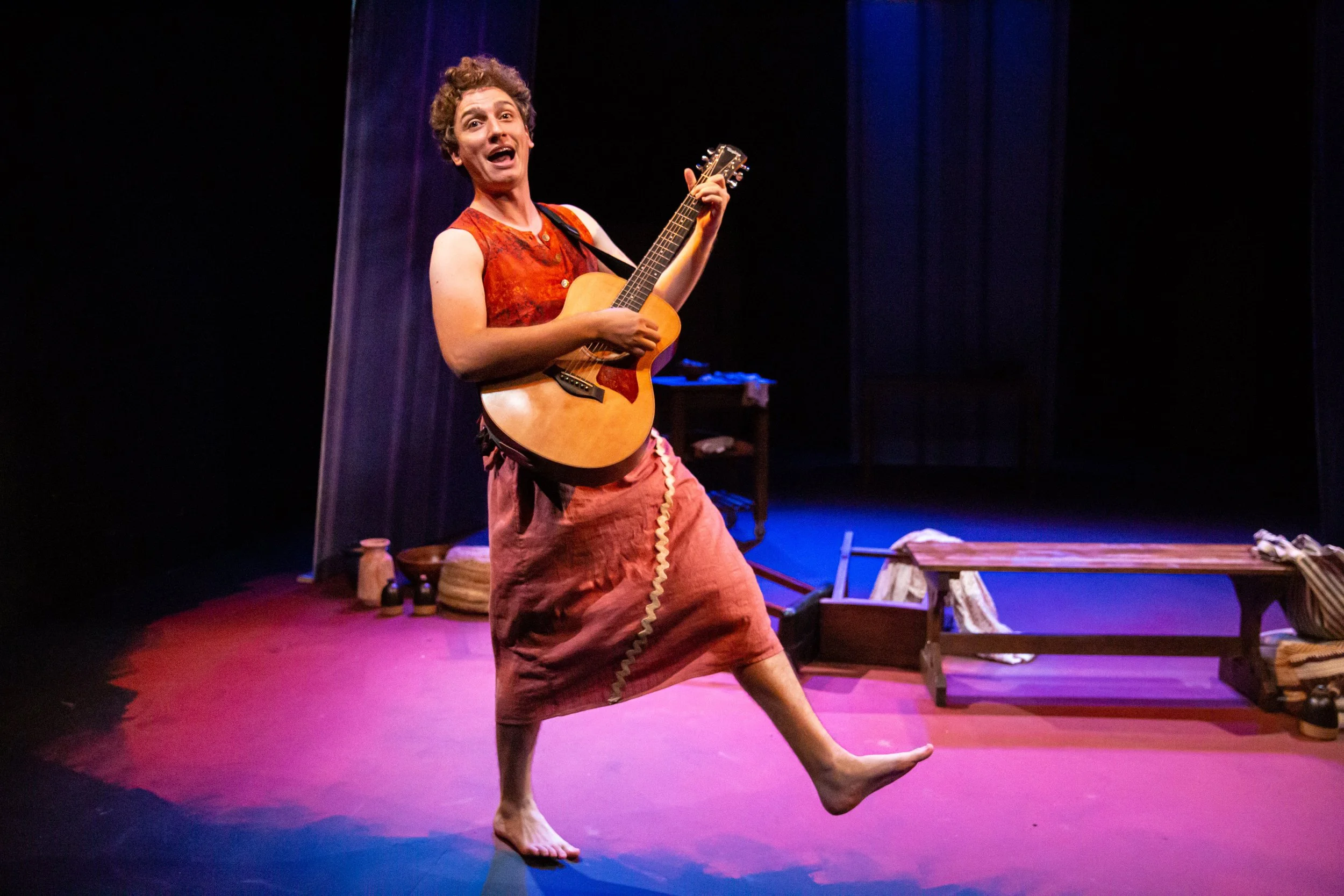 Performer playing a guitar on stage with theatrical set and props in the background.