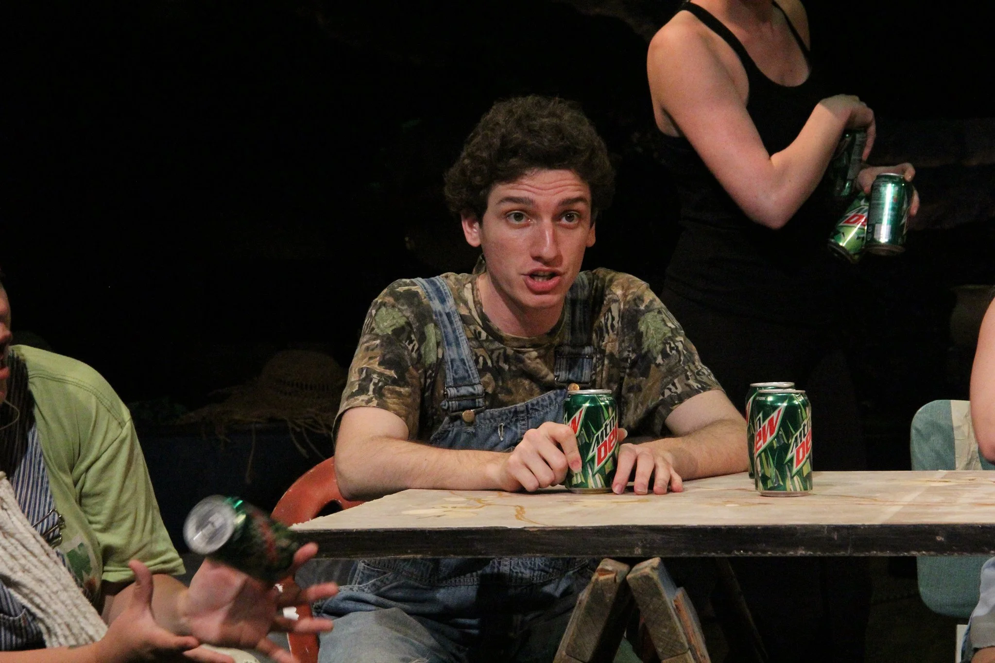 A young man with curly hair wearing camouflage shirt and denim overalls sitting at a table holding a can of Mountain Dew. There are more cans of Mountain Dew on the table. A woman next to him is visible holding a bottle. Another woman is in the backg