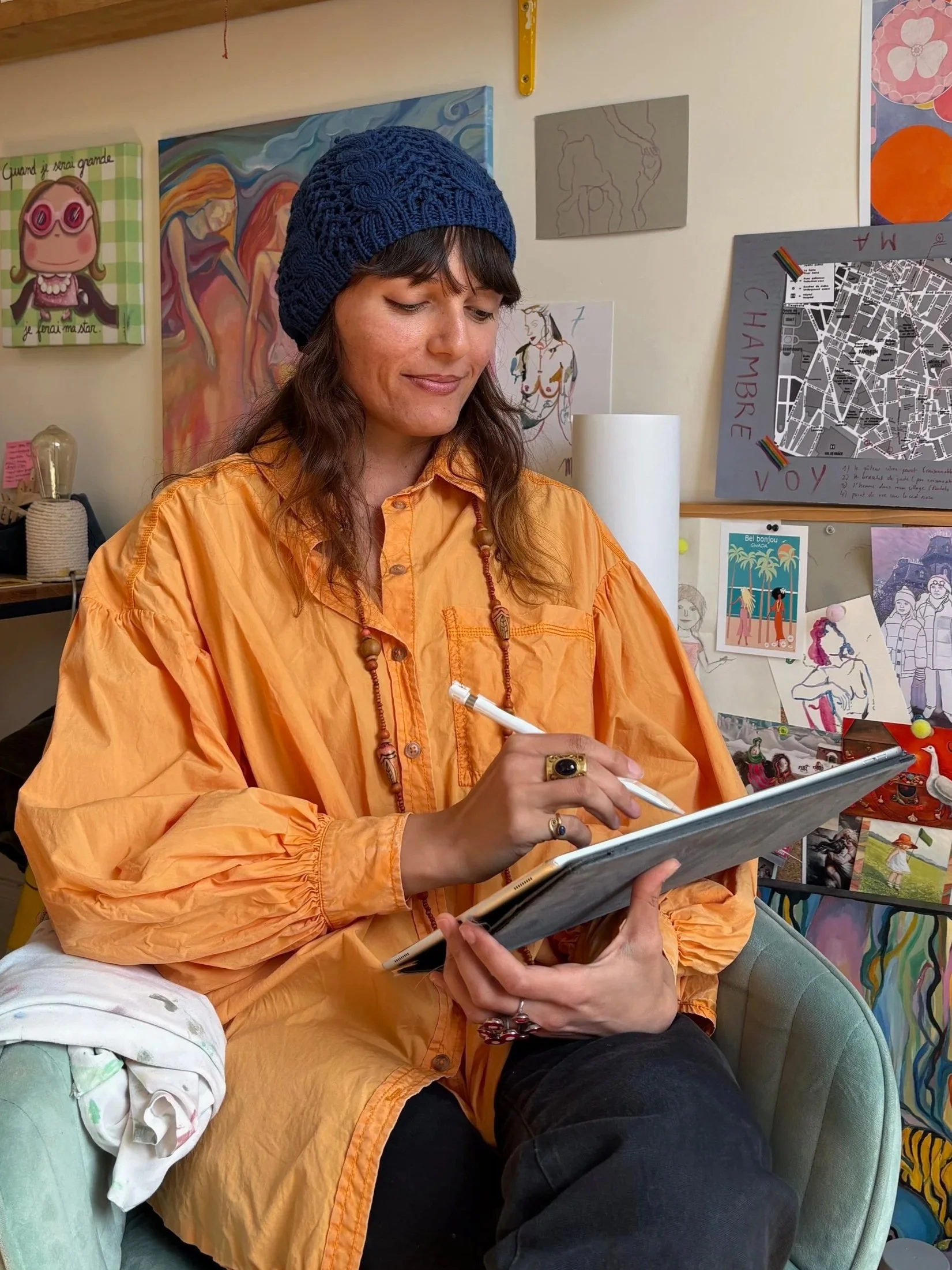 Une femme portant un bonnet bleu et une chemise orange, assise sur un fauteuil vert, peint sur une tablette numérique, dans un environnement décoré avec plusieurs œuvres d'art et posters muraux.