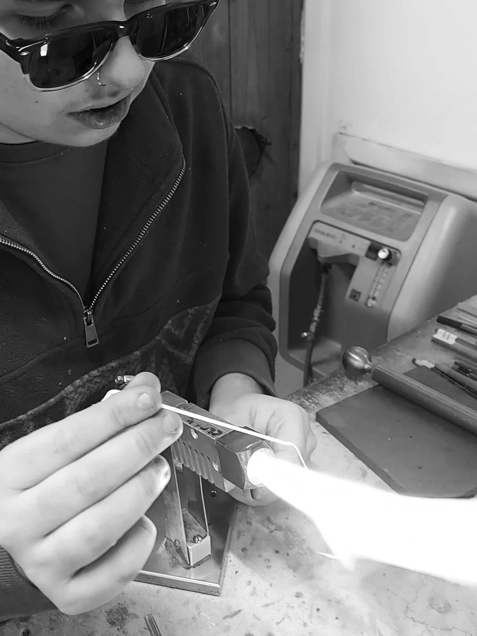 A person flameworking glass with metal and graphite tools, wearing diindium glasses, and using a torch at a workshop or studio.