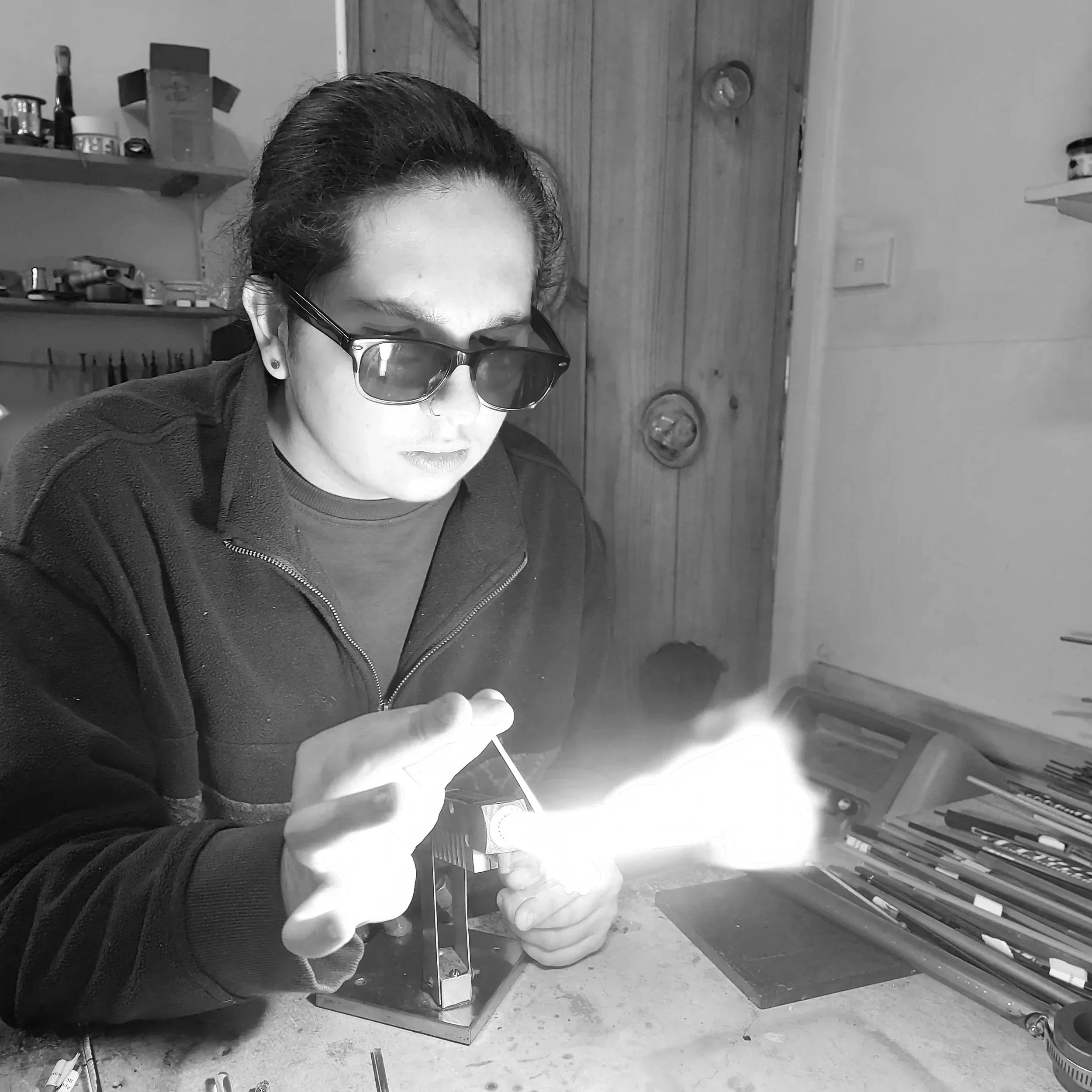 A person with short dark hair, wearing sunglasses and a dark jacket, is working on a metal object in a workshop. The person is holding a tool and appears to be welding or assembling the metal piece. The background shows shelves with various tools and materials, a wooden door, and a floor scattered with small items.