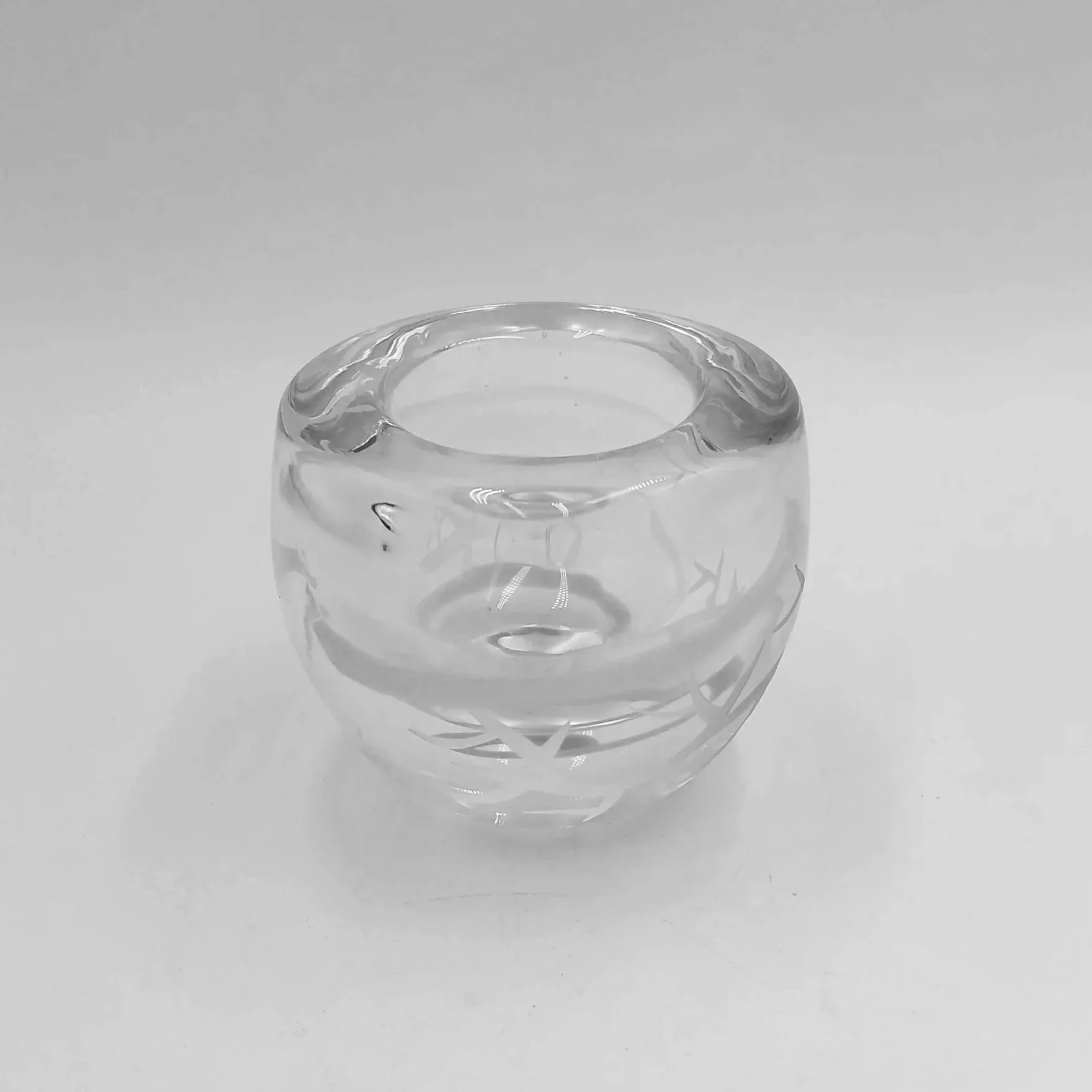 A clear, round glass vessel with a smooth surface, resting on a reflective surface with a light grey background.