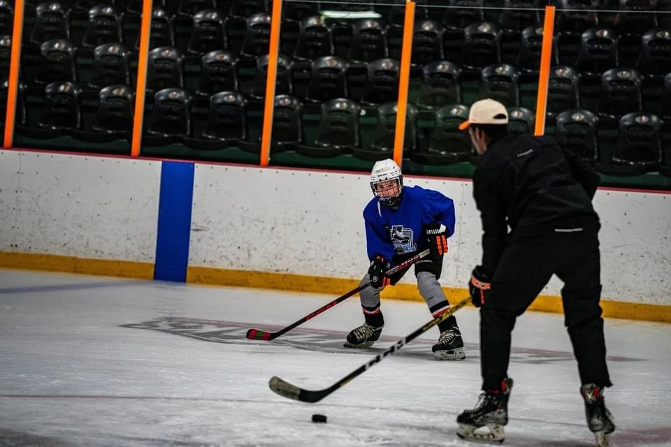A young hockey player practicing with a coach on an ice rink. The young player wears a blue jersey, helmet, and skates, while the coach is dressed in black with a white cap, coaching with a hockey stick. Empty bleacher seats are visible in the background.