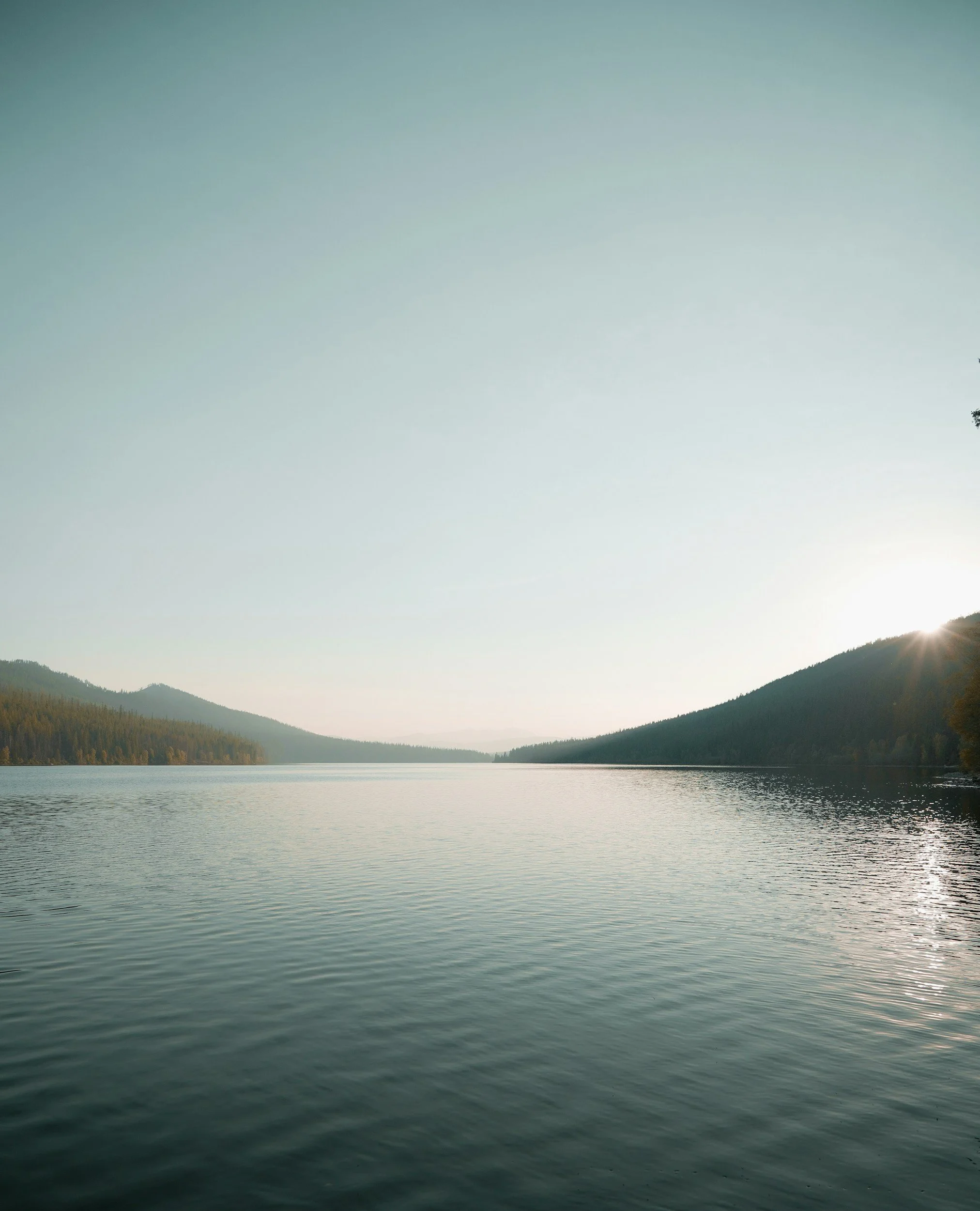 A large body of water, possibly a lake, surrounded by forested hills. The sky is clear with a gentle gradient from light to darker teal, and the sun is setting behind the hills on the right side of the image.