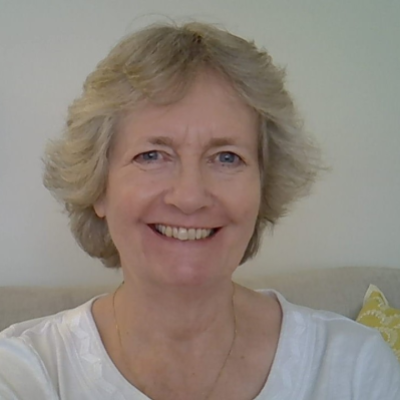 A smiling middle-aged woman with short, wavy blonde hair, wearing a white top, sitting in a room with a light-colored wall and a pillow in the background.