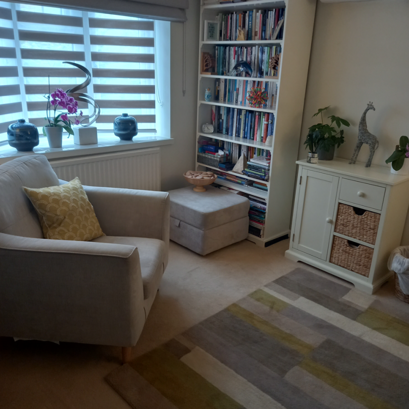 A cozy living room corner with a white armchair and a yellow patterned pillow, a window with striped blinds, decorative items on the windowsill, a tall bookshelf filled with books, a white cabinet with drawers and wicker baskets, and a beige ottoman with a bowl on top. There is a colorful striped rug on the floor and a houseplant with a giraffe figurine on the cabinet.