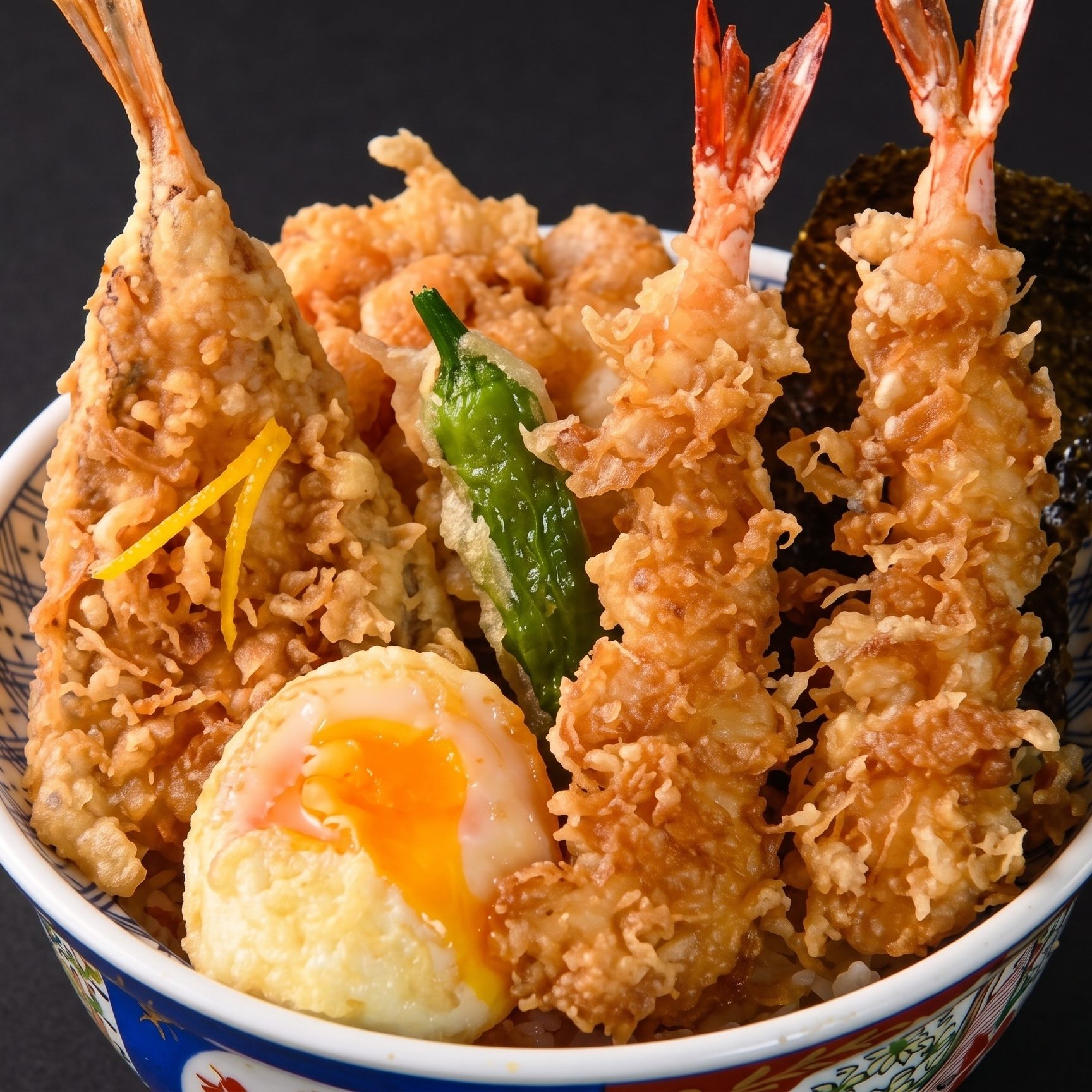 Close-up of a bowl of Japanese tempura shrimp with a soft-boiled egg with a runny yolk, green chili pepper, and shredded lemon peel, on a dark background.