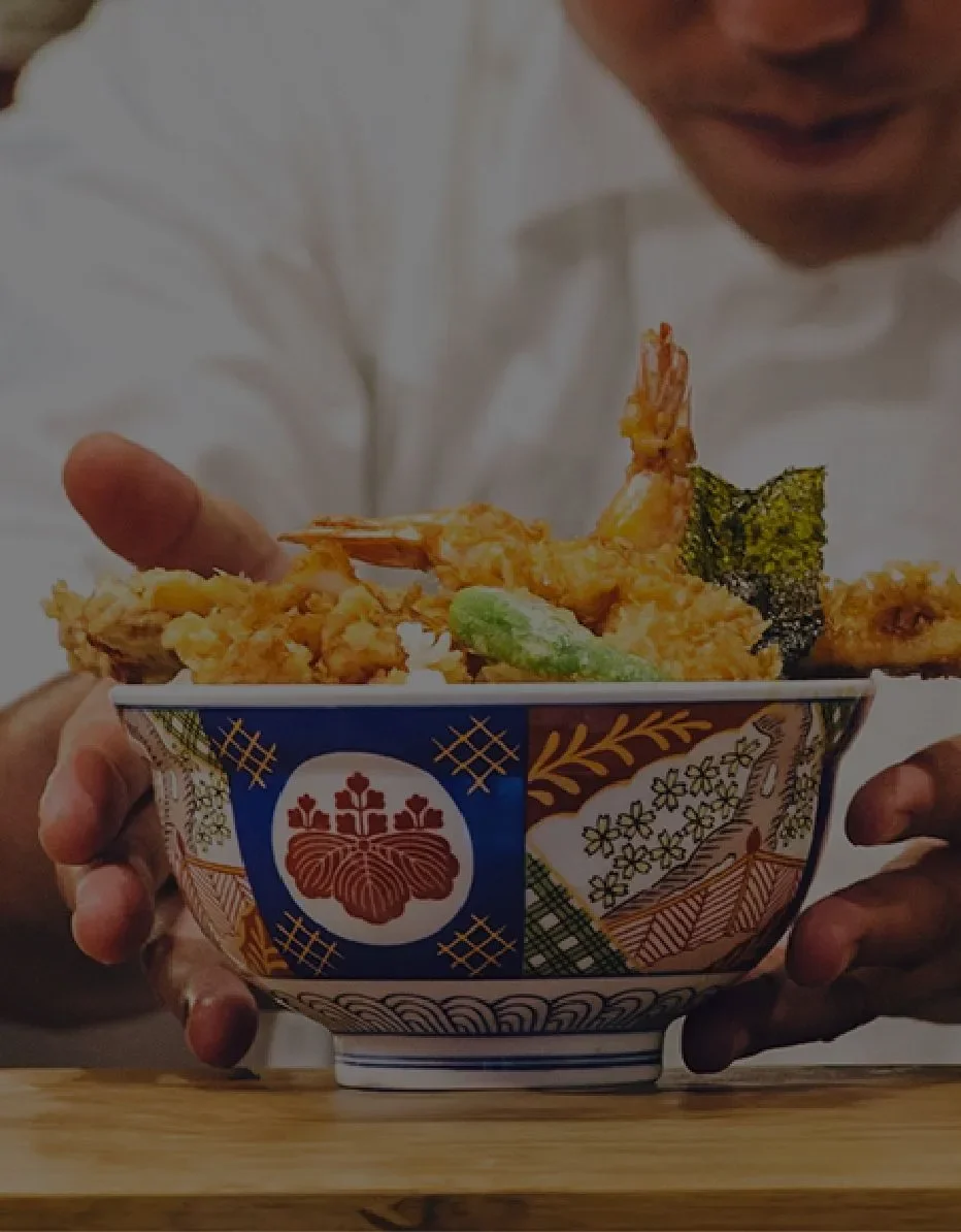 Person holding a bowl of tempura, including shrimp, fish, and vegetables, with traditional Japanese patterns on the bowl.