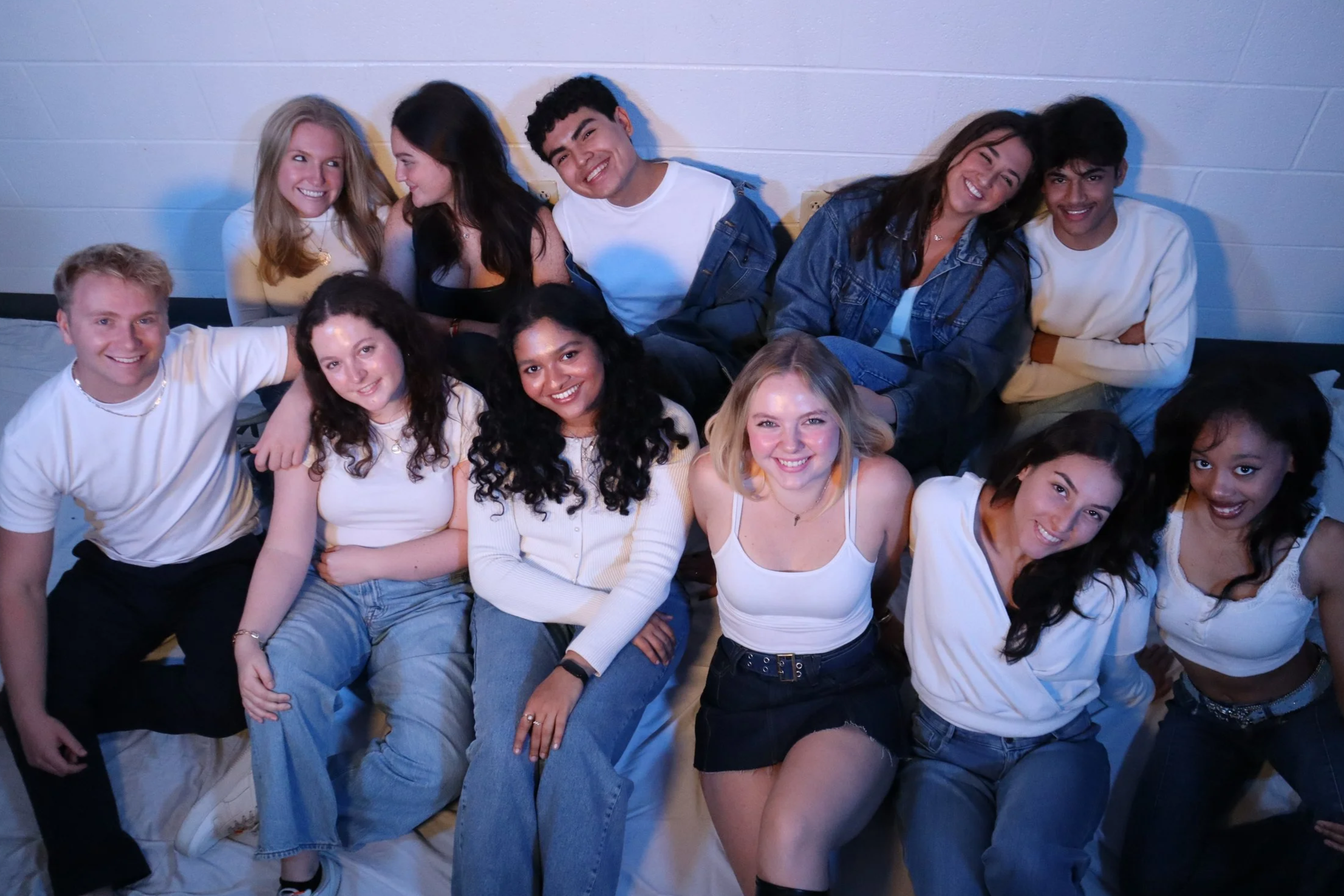 Group of diverse young people sitting and smiling against a white wall, wearing casual clothes, some in white shirts and jeans, in an indoor setting.