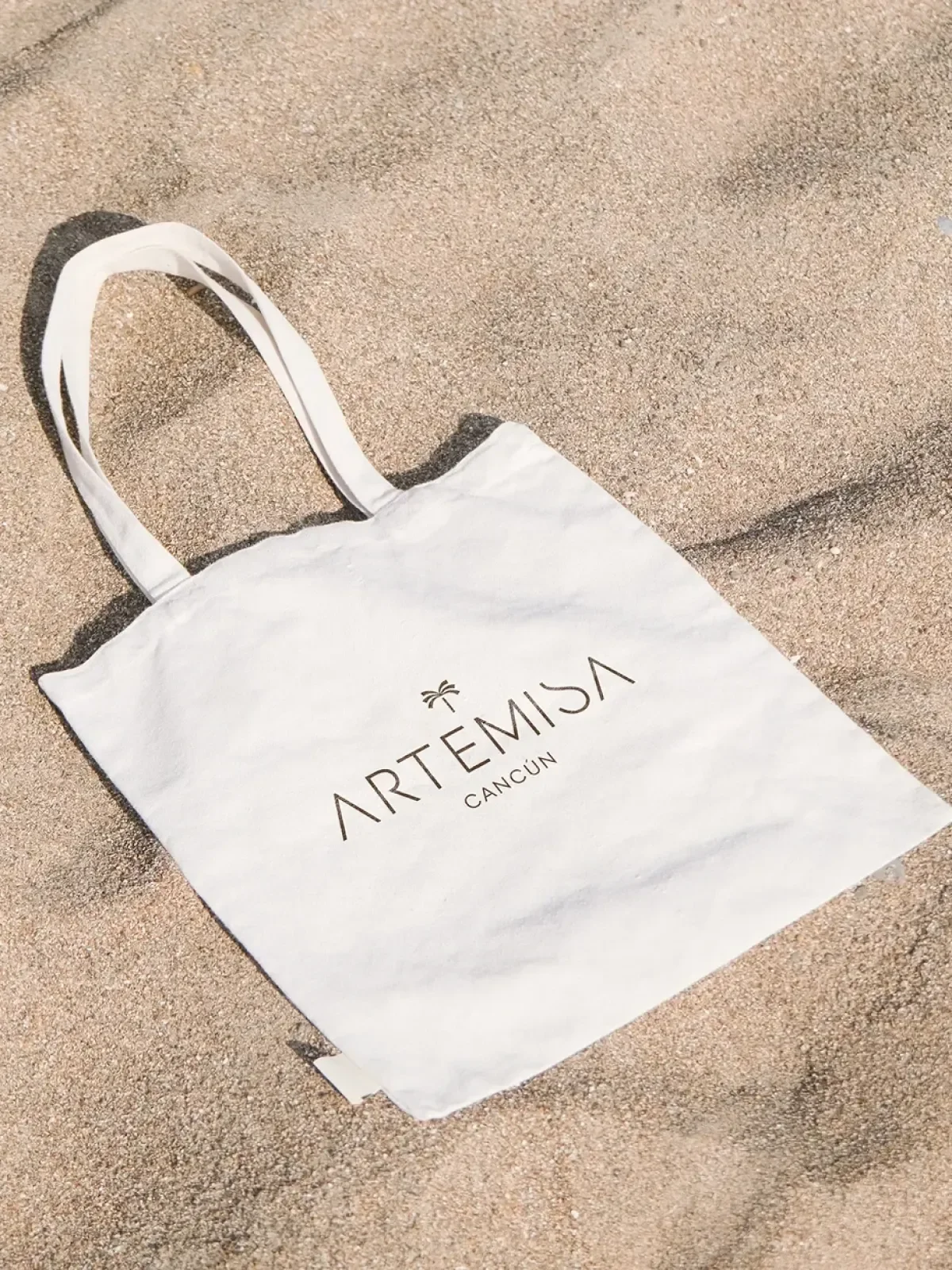 Tote bag de Artemisa Cancún sobre arena con diseño minimalista y branding del desarrollo