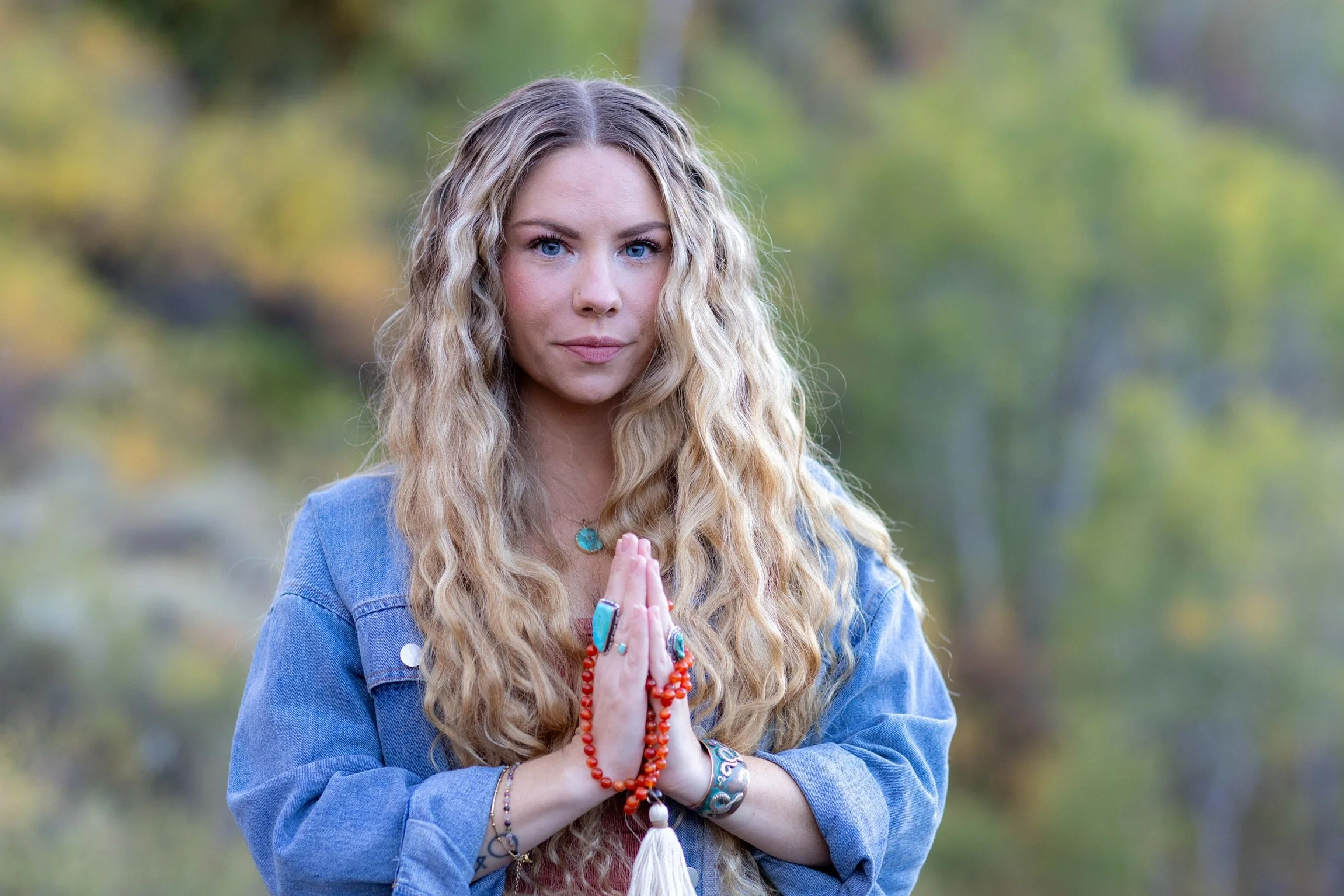 A woman with long curly blonde hair, blue eyes, wearing a denim jacket and colorful jewelry, standing outdoors with her hands pressed together in a prayer or meditation pose.