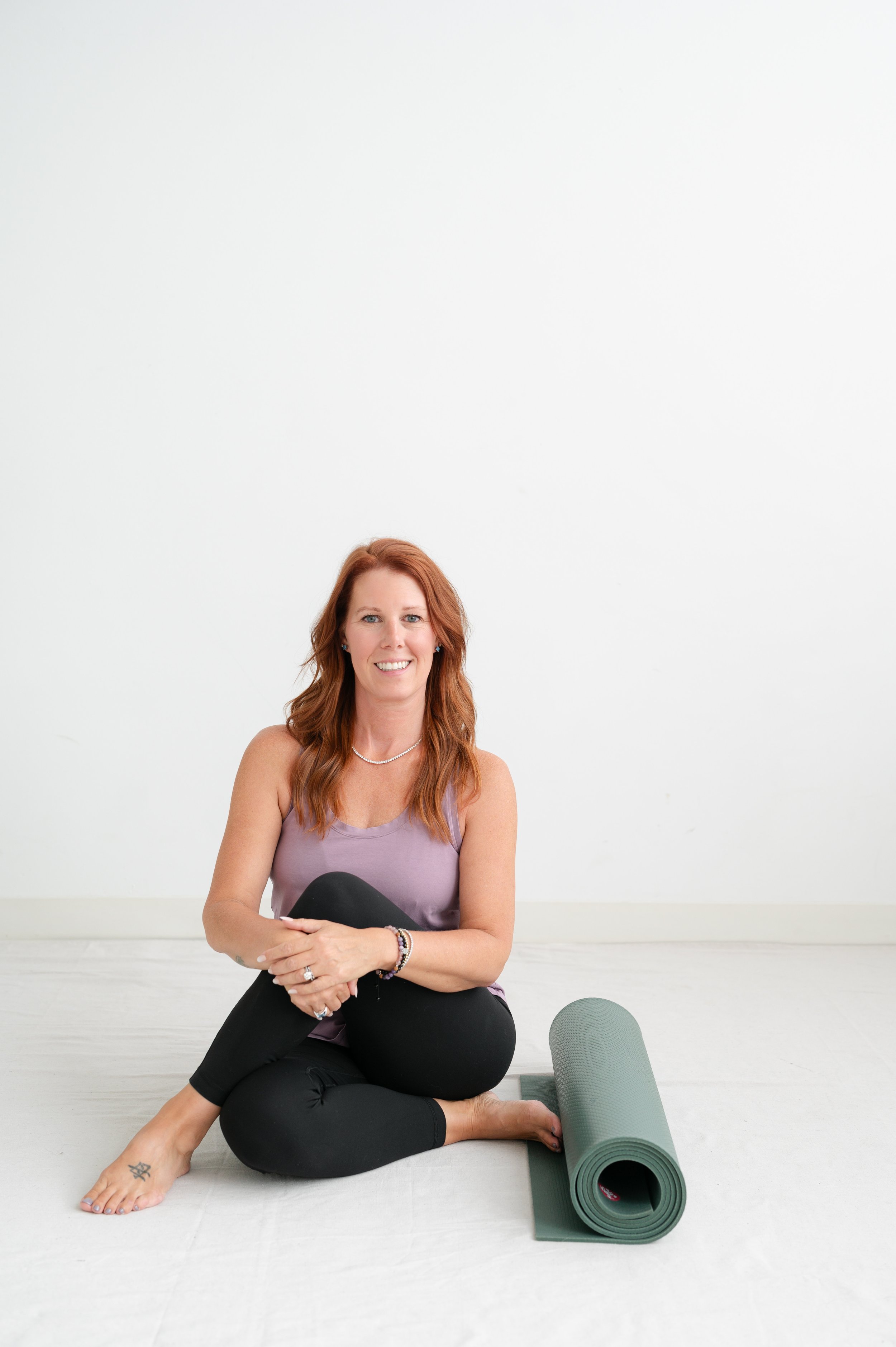 A woman with long red hair and tattoos, sitting on the floor with crossed legs, smiling, wearing a lavender tank top and black pants, next to a rolled-up green yoga mat in a bright, minimalistic room.