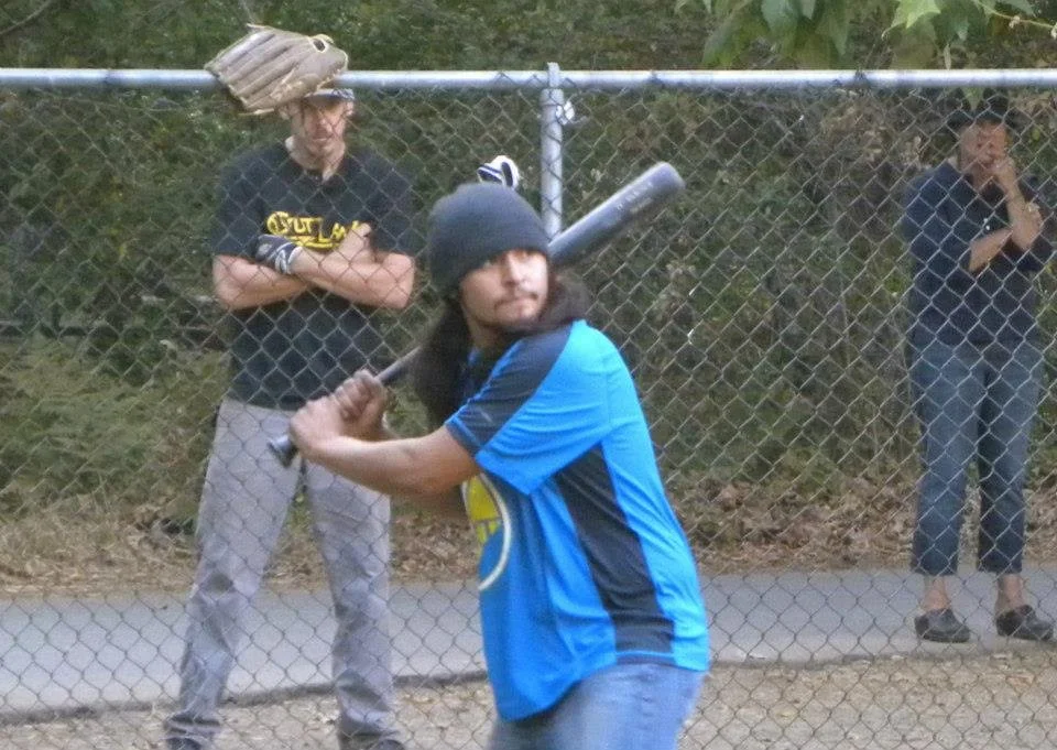 A person in a blue and black shirt holding a baseball bat, standing behind a chain-link fence. Two other people are in the background, one with folded arms and the other with a hand on their face, observing