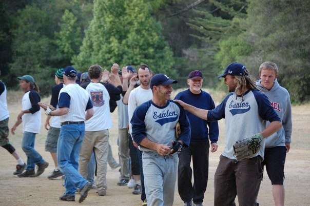 A group of people, including baseball players in Saalen jerseys, gather outdoors on a dirt field with trees in the background. Some are talking, and one person is high-fiving another.