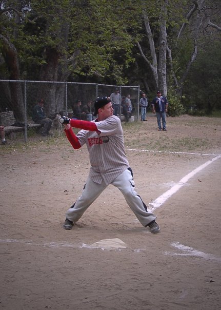 A young baseball player at bat during a game, standing on home plate in a grey uniform, holding a bat, with a chain-link fence and spectators in the background.