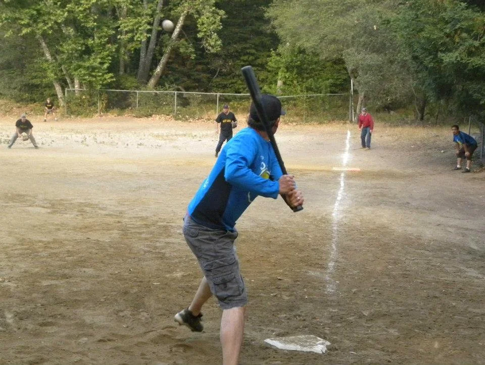 Person in blue jacket preparing to swing a baseball bat at a softball on a dirt field, with other players and trees in the background.