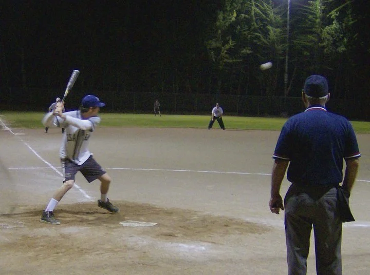 A young baseball player at bat during a game, with an adult umpire behind him and other players in the outfield at night on a baseball field.