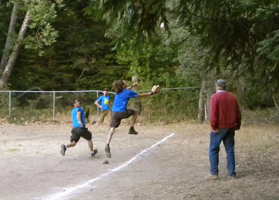 Children playing baseball, with one child jumping to catch the ball while another runs towards the baseball diamond's white line, and an adult nearby watching, in a wooded outdoor setting.