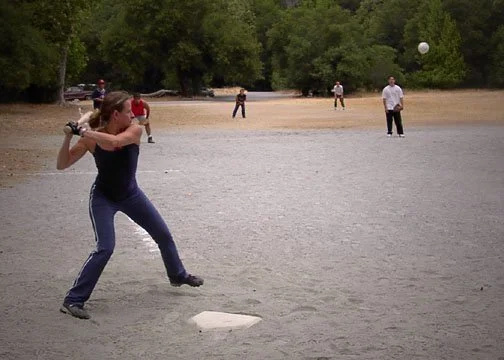 A woman swinging a baseball bat at a baseball on a field with other people in the background.