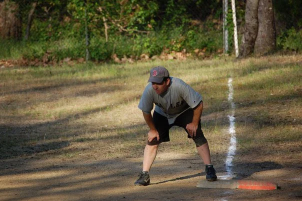 A young man wearing a gray baseball cap, gray t-shirt, and black shorts is in a bent-over stance on a sports field, near a marked line and a base, preparing for a baseball or softball game.