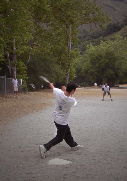 Child swinging a baseball bat at a baseball game in a park, with other players and tall trees in the background.