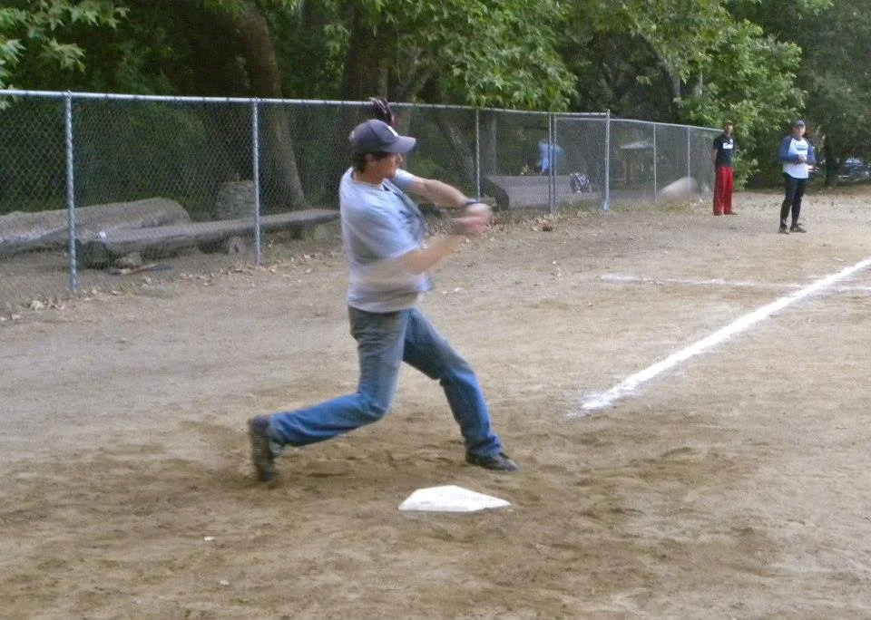 A woman in a baseball cap and casual clothes is swinging a bat at a baseball on a dirt field during daytime.
