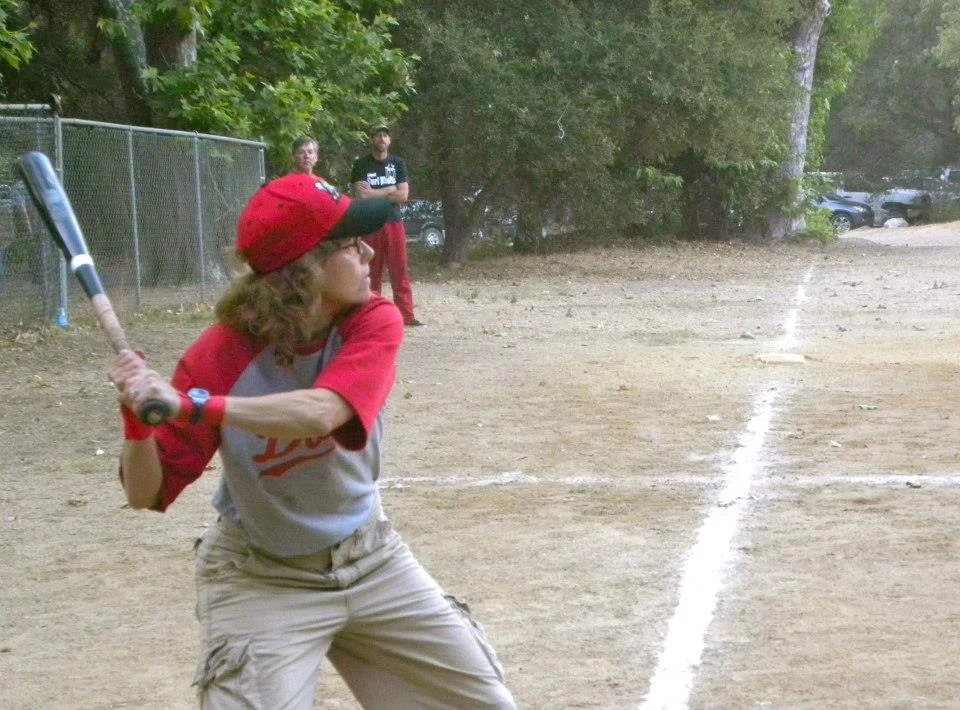 A young boy with long hair, wearing a red cap, glasses, a gray and red baseball jersey, and khaki pants, is in batting stance on a baseball field, holding a bat ready to hit a pitch.
