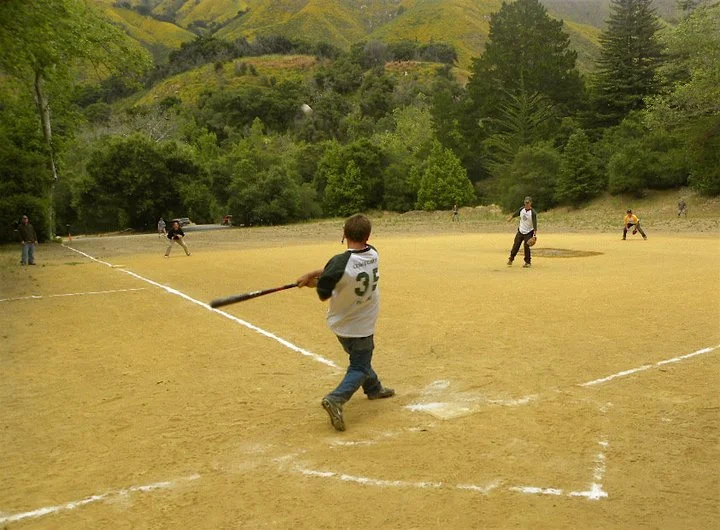 Kids playing baseball on a dirt field surrounded by trees and hills in the background.