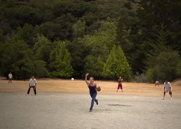 People playing baseball outdoors on a field with trees in the background.